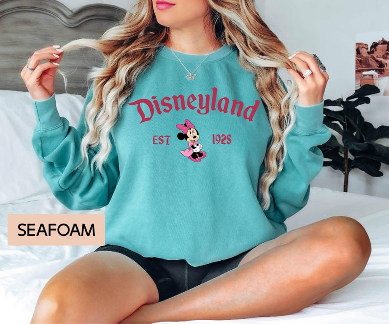 May include: Seafoam green sweatshirt with the word "Disneyland" in red script, the year "EST 1928", and a Minnie Mouse graphic. The sweatshirt is a crew neck style.