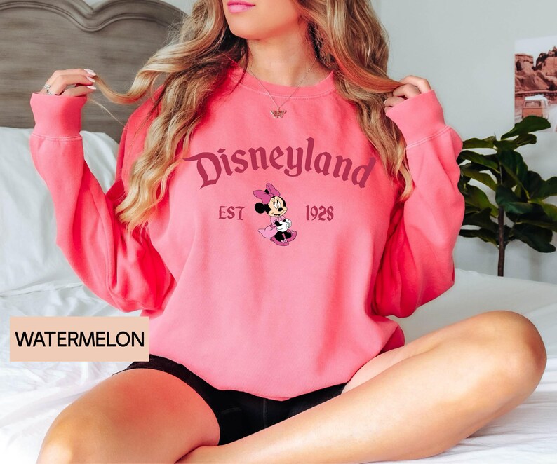 May include: A watermelon-colored sweatshirt featuring the Disneyland logo with Minnie Mouse. The design includes the text "Disneyland EST 1928". The sweatshirt has long sleeves and a crew neck.