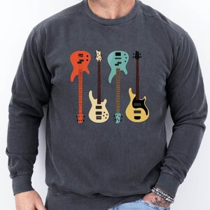May include: Dark gray sweatshirt featuring a colorful graphic of four guitars in shades of red, cream, teal, and yellow. The guitars are arranged vertically, with a vintage-inspired design. The sweatshirt is a crew neck style.