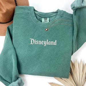 May include: A green sweatshirt with the word "Disneyland" embroidered on the front in white. The sweatshirt has a distressed look and is made of a soft, comfortable fabric.