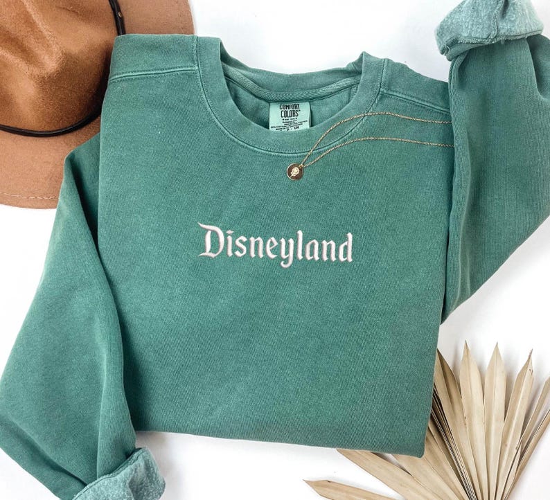 May include: A green sweatshirt with the word "Disneyland" embroidered on the front in white. The sweatshirt has a distressed look and is made of a soft, comfortable fabric.