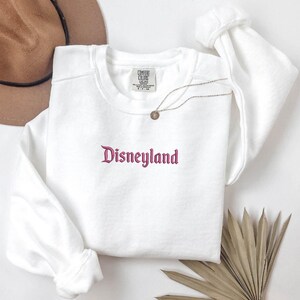 May include: White sweatshirt with a pink embroidered Disneyland logo. The sweatshirt has a crew neck and long sleeves.