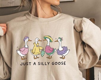 May include: A beige sweatshirt featuring a whimsical illustration of four geese dressed in various outfits, including hats and dresses. The text "JUST A SILLY GOOSE" is printed below the geese. The sweatshirt is a casual, comfortable style.
