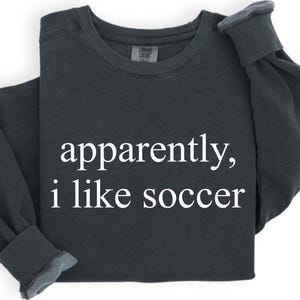 May include: Dark gray sweatshirt with the text "apparently, i like soccer" in white. The long-sleeved shirt has a crew neck and ribbed cuffs. The garment is folded, showing the text prominently.