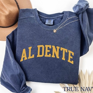May include: A navy blue sweatshirt with the text "AL DENTE" in yellow, printed in an arch across the front. The sweatshirt is made of a soft, comfortable fabric and has a relaxed fit. The text "TRUE NAVY" is printed in white on the bottom right corner of the sweatshirt.