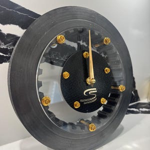 Puede incluir: Un reloj único con una esfera de fibra de carbono negra y un marco de acrílico transparente. El reloj presenta tornillos y manecillas doradas, y un centro negro con un logotipo plateado. El diseño del reloj es industrial y moderno.