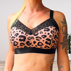 May include: A bralette featuring a peach-colored fabric with a black leopard print pattern. The garment includes black lace trim along the neckline and a wide, solid black elastic band at the base, complemented by thick black shoulder straps.