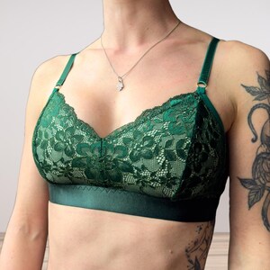 May include: A dark green lace bra with adjustable straps and a satin band. The bra features a floral lace pattern and a delicate design. The straps are attached with gold-colored metal rings.