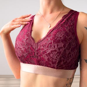 May include: A deep burgundy lace bralette featuring a floral pattern throughout the fabric, a V-neckline, and a wide, smooth, light pink elastic band at the base. The garment has thick shoulder straps and a structured design with visible seams.