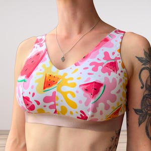 Può includere: Reggiseno sportivo bianco, rosa e giallo con motivo anguria. Il reggiseno presenta uno scollo a V e una fascia color beige chiaro. Le fette di anguria e gli schizzi di colore creano un'atmosfera divertente ed estiva.