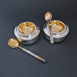 Può includere: Due ciotole da portata argento e oro con cucchiai abbinati. Le ciotole hanno un esterno argentato e un interno dorato. I cucchiai hanno teste di cucchiaio dorate e manici argentati. Gli oggetti sono su sfondo scuro.