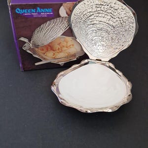 Könnte beinhalten: Eine silberfarbene, muschelförmige Servierschale mit weißem Innenraum. Die Schale ist geöffnet und zeigt den weißen Innenraum und die strukturierte Außenseite. Auf der Schachtel im Hintergrund steht "Queen Anne, A Mayell Product, Made in England."