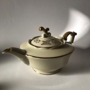 Può includere: Una teiera bianca con un bordo dorato e un motivo floreale. La teiera ha un coperchio con un manico dorato e un pomo dorato.