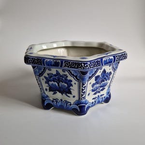 Può includere: Un vaso di ceramica blu e bianco a forma esagonale. Il vaso presenta un motivo floreale blu su sfondo bianco. Ha piedini e un bordo decorativo. Il vaso misura circa 20 cm di larghezza.