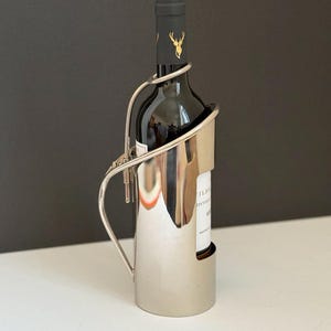 Può includere: Un portabottiglie d'argento per vino culla una bottiglia di vino scuro. Il supporto ha un design elegante e moderno con un manico curvo e una base che sostiene la bottiglia. La bottiglia di vino ha un'etichetta nera con dettagli dorati.