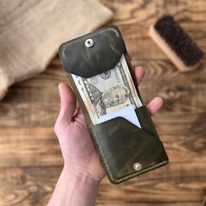 May include: A dark green leather wallet with a snap closure, holding several banknotes and a white card. The wallet has a minimalist design and appears to be handmade. The leather has a distressed finish.