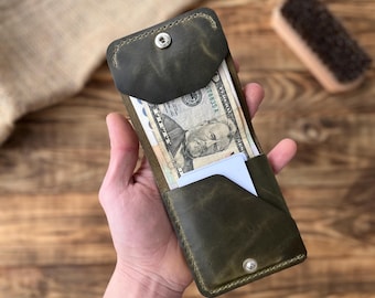 May include: A dark green leather wallet with a snap closure, holding several U.S. dollar bills and a white card. The wallet has a minimalist design and appears to be handmade. The leather has a distressed finish.