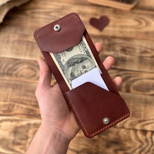 May include: A burgundy leather wallet with a snap closure, holding a one-hundred-dollar bill and a white card. The wallet is rectangular and has a stitched border. The background is a wooden surface.