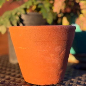 May include: A terracotta plant pot with a matte, porous surface and a warm orange-brown hue. The vessel features a simple, tapered cylindrical shape with a flat rim and a slightly textured exterior, showcasing a classic earthy appearance.