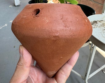 May include: A terracotta-colored, diamond-shaped clay vessel with a pointed base and a flat top. The object features a single circular hole near the upper edge, showcasing a matte, earthy texture and a distinct geometric form with angled sides.