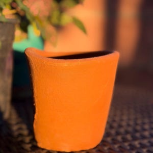May include: A terracotta plant pot with a matte, orange-brown finish and a slightly uneven, wavy rim. The vessel has a tapered cylindrical shape with a smooth, porous texture, showcasing a simple, unadorned design in a warm, earthy tone.