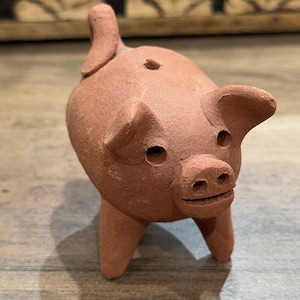 Puede incluir: Una pequeña figurita de cerdo de terracota con la cola enroscada, de pie sobre cuatro patas. El cerdo tiene un cuerpo redondo, un hocico y dos orejas pequeñas.