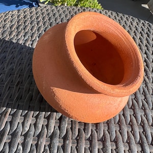 Puede incluir: Una vasija de barro de color terracota con una boca ancha y abierta y un borde grueso y texturizado. La superficie exterior del recipiente tiene un acabado mate y poroso con un tono marrón anaranjado, mostrando un diseño simple y abierto.