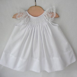 May include: White baby dress with lace trim on the sleeves. The dress has a smocked bodice and a full skirt.