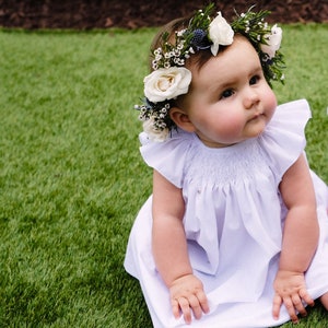 May include: A baby girl wearing a white dress with a flower crown on her head. The crown is made of white flowers and green leaves. The baby is sitting on a green grassy surface.