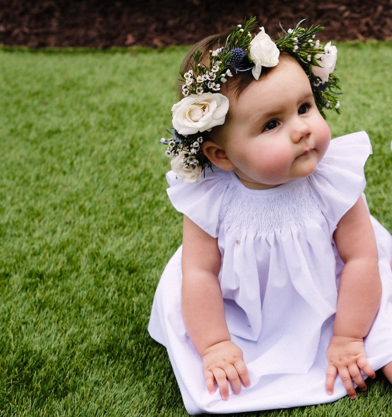 May include: A baby girl wearing a white dress with a flower crown on her head. The crown is made of white flowers and green leaves. The baby is sitting on a green grassy surface.