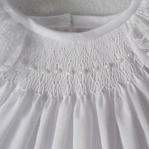 May include: White baby dress with a smocked bodice and lace trim on the sleeves. The bodice is decorated with a row of pearl beads.
