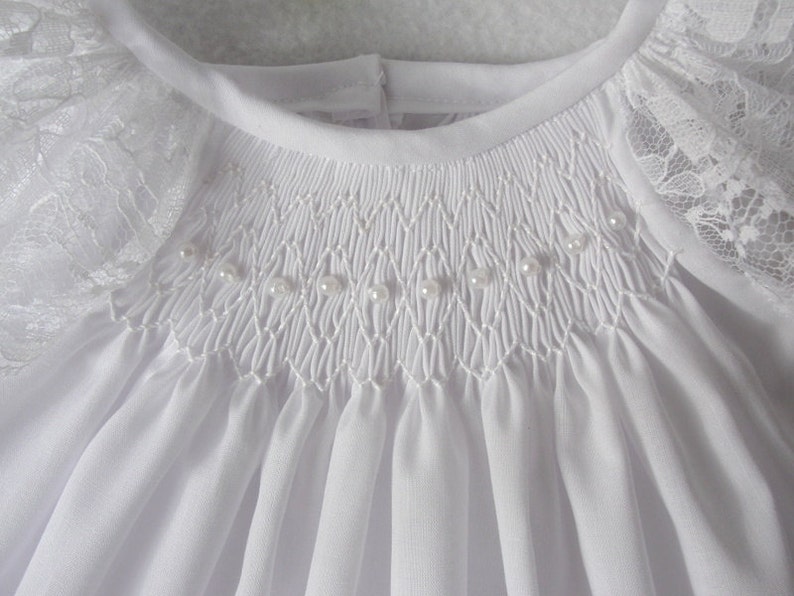 May include: White baby dress with a smocked bodice and lace trim on the sleeves. The bodice is decorated with a row of pearl beads.