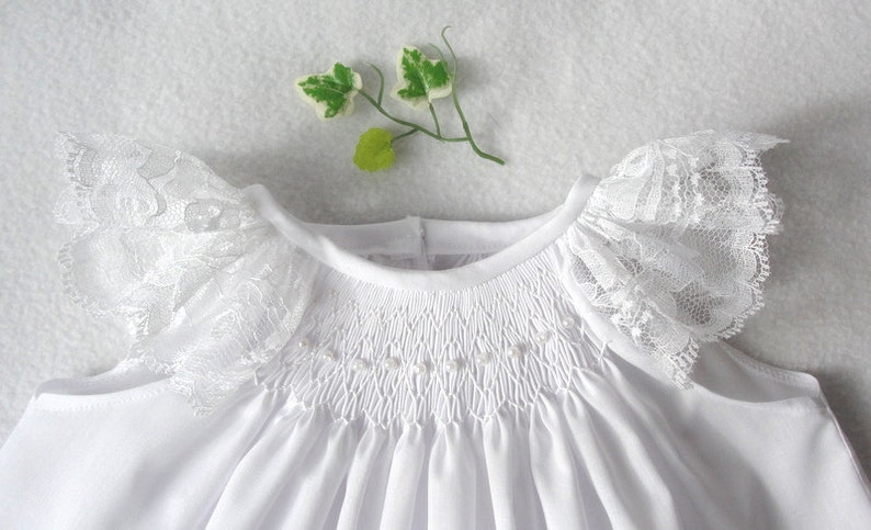 May include: White baby dress with a smocked bodice and lace trim on the sleeves.
