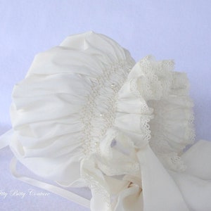 May include: A white baby bonnet with a lace trim and pearl embellishments. The bonnet has a satin ribbon tied at the back.