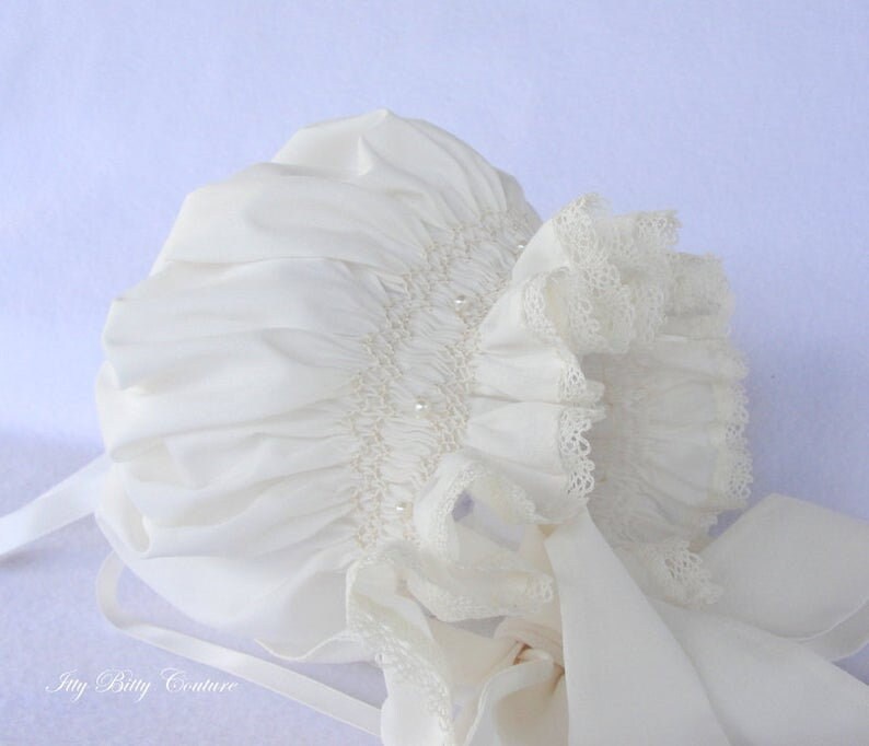 May include: A white baby bonnet with a lace trim and pearl embellishments. The bonnet has a satin ribbon tied at the back.