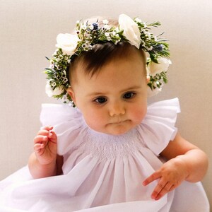May include: A baby girl wearing a white dress with a flower crown made of white flowers and green leaves.
