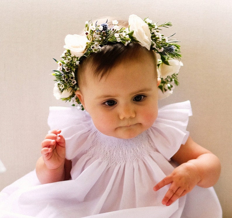 May include: A baby girl wearing a white dress with a flower crown made of white flowers and green leaves.