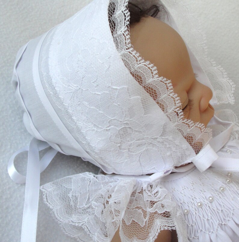 May include: White lace baby bonnet with a satin ribbon tie. The bonnet has a delicate floral pattern and is perfect for a christening or special occasion.