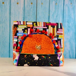 May include: A colourful fabric clutch with a fuzzy orange flap and a gold cat charm. The clutch has a black background with a pattern of colourful books and a second flap with a black background and a pattern of white rabbits and carrots on a black background.