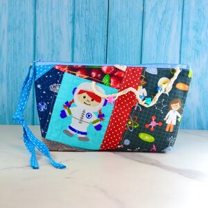 Peut inclure: Une pochette zippée bleu et blanc avec un motif patchwork représentant une petite fille astronaute, des cerises et des motifs sur le thème des sciences. La pochette est dotée d'un cordon de serrage bleu et blanc à pois.