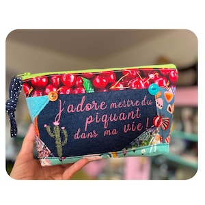 May include: A zipped pouch featuring a cherry print top, a dark blue centre panel with the embroidered text J'adore mettre du piquant dans ma vie!, a cactus graphic, and colourful buttons. The pouch has a lime green zip and a navy polka dot pull.