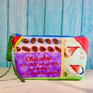 May include: A colourful zippered pouch with a patchwork design featuring chocolate squares, gnomes, and the text "Chocolat" in French. The pouch has a green and brown strap.