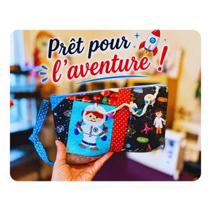May include: A rectangular fabric zip pouch featuring a patchwork design with an astronaut character, red polka dots, and a black space-themed print with science icons. The text Prêt pour l'aventure! and a rocket ship graphic appear above.