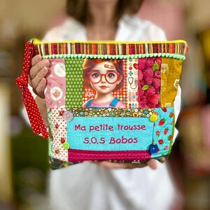 Peut inclure: Une trousse patchwork colorée avec une fermeture éclair et une poignée à pois rouges. L'avant présente une image de dessin animé d'une fille portant des lunettes et le texte "Ma petite trousse S.O.S Bobos".