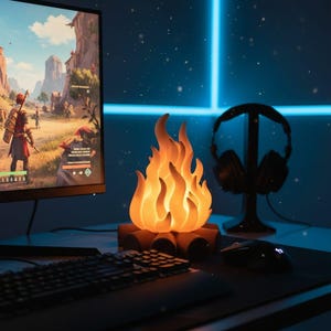 May include: A decorative campfire-shaped lamp glows on a desk beside a computer monitor showing a video game. A black keyboard, mouse, and headset stand are also present. The lamp gives off a warm orange light, creating a cosy gaming setup.