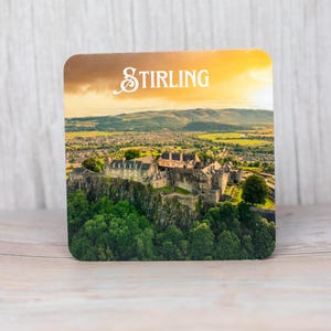 Könnte beinhalten: Quadratischer Untersetzer mit einem Foto von Stirling Castle in Schottland, mit dem Wort "Stirling" in Weiß oben. Das Bild zeigt das Schloss auf einem grünen Hügel mit einem Sonnenuntergangshimmel.