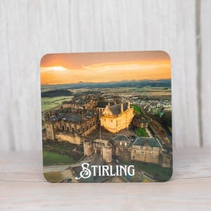 Könnte beinhalten: Quadratischer Untersetzer mit einer Luftaufnahme von Stirling Castle in Schottland, mit dem Wort "STIRLING" in weißer Schrift. Das Bild zeigt das Schloss bei Sonnenuntergang mit einem leuchtend orange-gelben Himmel.