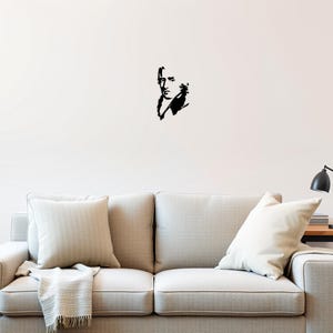 May include: Black and white silhouette of a man's face with a serious expression. The silhouette is on a white wall above a light beige sofa with two cushions.