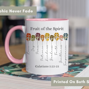 May include: White ceramic mug with a pink interior and handle. The mug features the text "Fruit of the Spirit" and the words "Love, Joy, Peace, Patience, Kindness, Goodness, Faith, Gentleness, Temperance" with floral illustrations. The text "Galatians 5:22-23" is also printed on the mug.