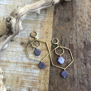May include: A pair of gold geometric earrings with purple gemstone accents. The earrings feature two hexagonal frames with a smaller hexagon inside each frame. The gemstones dangle from the bottom of the smaller hexagons.